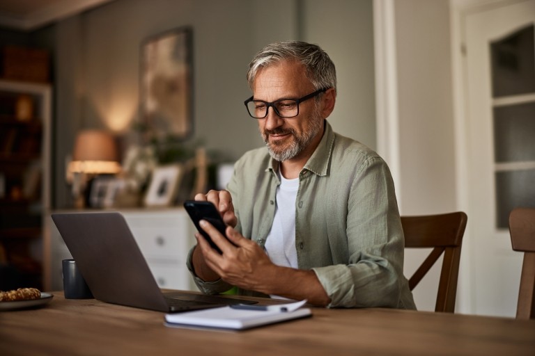 man working at a table with a laptop in front of him and looking at his cellphone in his hands